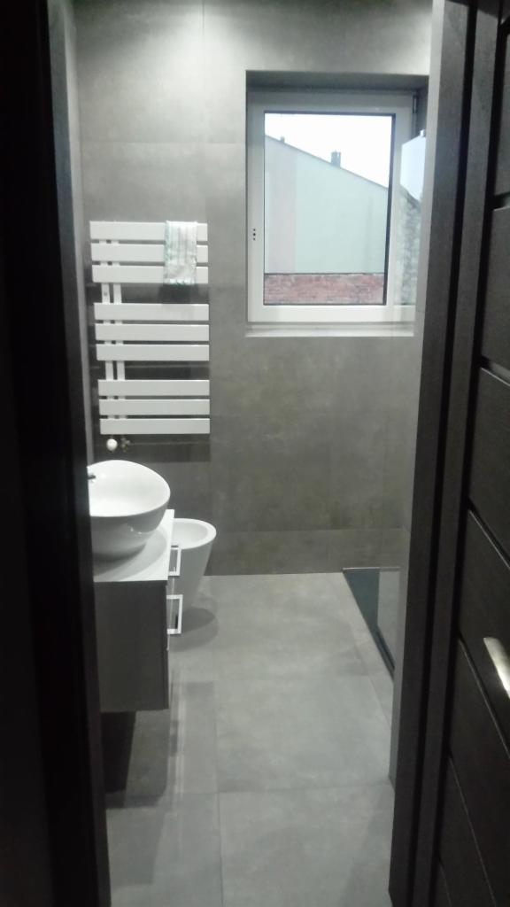 Nowoczesna, szara łazienka z grzejnikiem drabinkowym, umywalką nablatową i widokiem z okna na budynek z ceglanym dachem, widziana zza uchylonych drzwi.