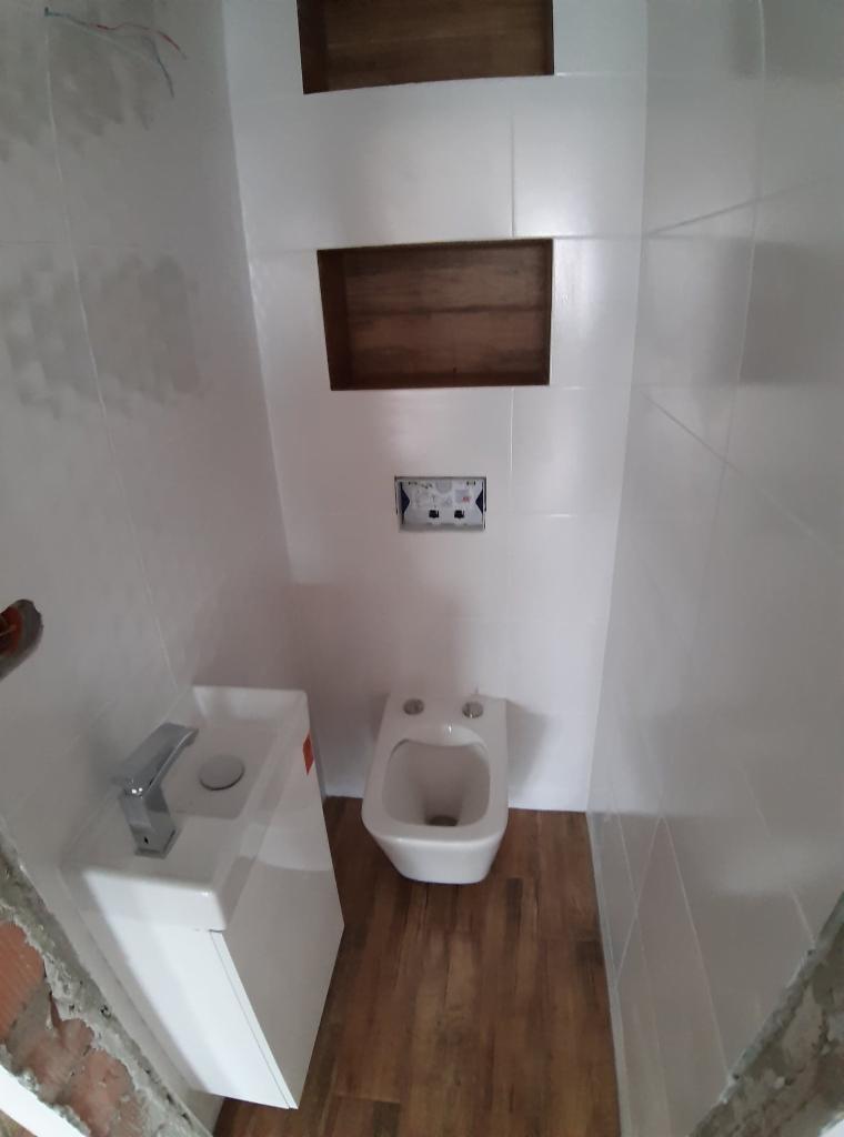 Wnętrze łazienki w trakcie wykańczania: białe płytki, drewnopodobna podłoga, zabudowana umywalka z chromowaną baterią, bidet i wnęki w ścianie z drewnianym wykończeniem, widoczne ślady po...