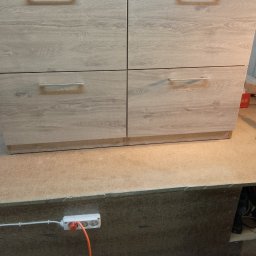 Meblesystem - Cztery szuflady z jasnego drewna z poziomymi, srebrnymi uchwytami, umieszczone na blacie roboczym. Widoczny fragment przedłużacza i ciemne tło pod blatem.