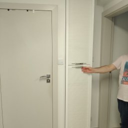 Meblesystem - Biała szafa wnękowa z żaluzjowymi drzwiami, otwierana przez osobę. Widoczna obok biała ściana z włącznikiem i białe drzwi. Minimalistyczny design.