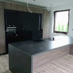 Meblesystem - Nowoczesna kuchnia z wyspą: szare fronty szafek, czarny panel z piekarnikiem i mikrofalówką, minimalistyczny design, surowy stan wykończenia.