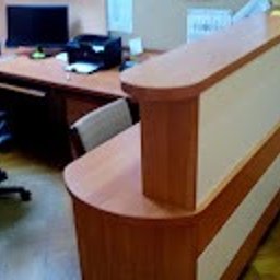 Meblesystem - Recepcja z blatem w odcieniach drewna i białymi panelami, widoczna z boku, z biurkiem i sprzętem biurowym w tle. Ujęcie z niskiej perspektywy.