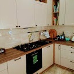 Meblesystem - Jasna kuchnia w kształcie litery L z drewnianym blatem i czarną płytą grzewczą. Na półkach widoczne książki i akcesoria kuchenne. Ujęcie z bliska, naturalne światło.