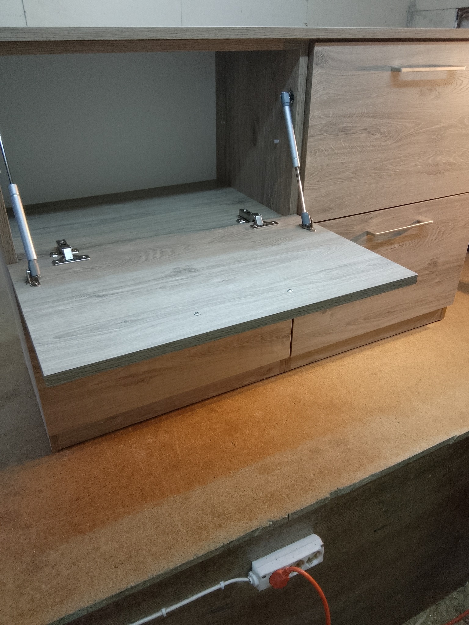 Otwarta szafka z mechanizmem podnoszenia frontu, widoczne zawiasy i siłowniki. Jasne drewno, minimalistyczny design, szuflady pod spodem. Detal meblowy w warsztacie.