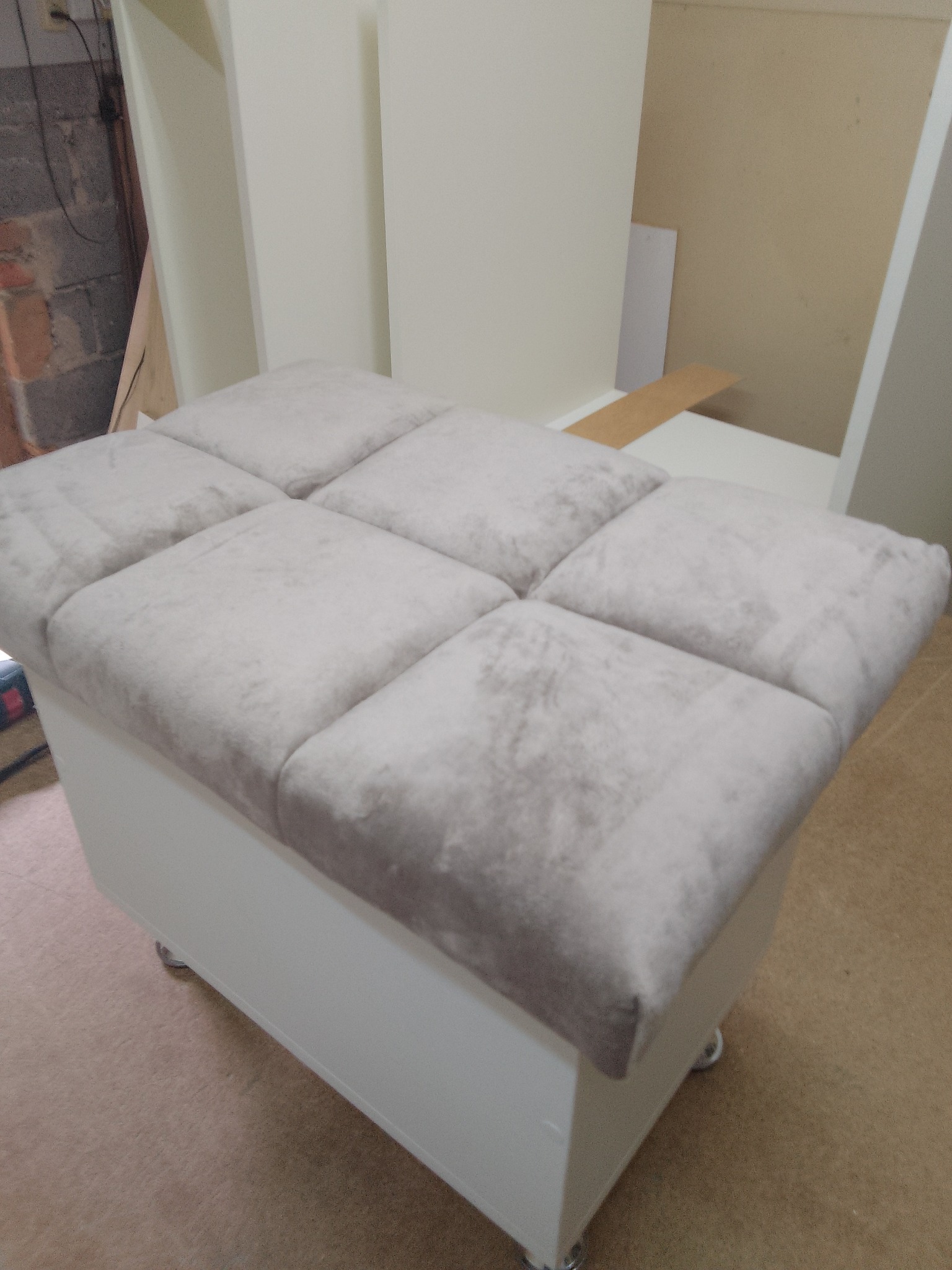 Biała pufa na kółkach z tapicerowanym, kwadratowym siedziskiem w odcieniach szarości. Widoczne fragmenty ściany z cegły i elementów meblowych w tle.