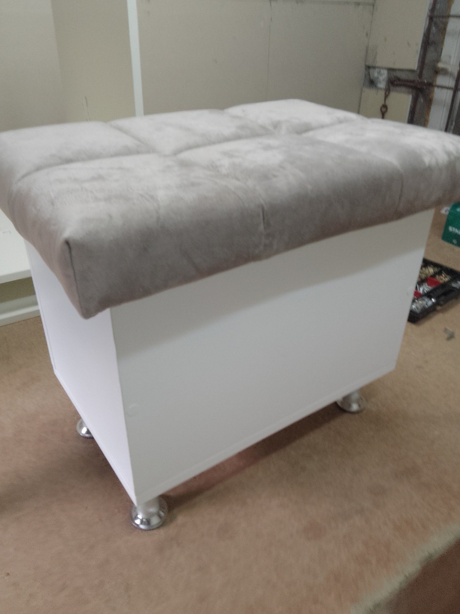 Tapicerowany puf z białym korpusem i srebrnymi nóżkami. Mebel z miękkim siedziskiem w odcieniach szarości, widoczny na tle warsztatu stolarskiego.