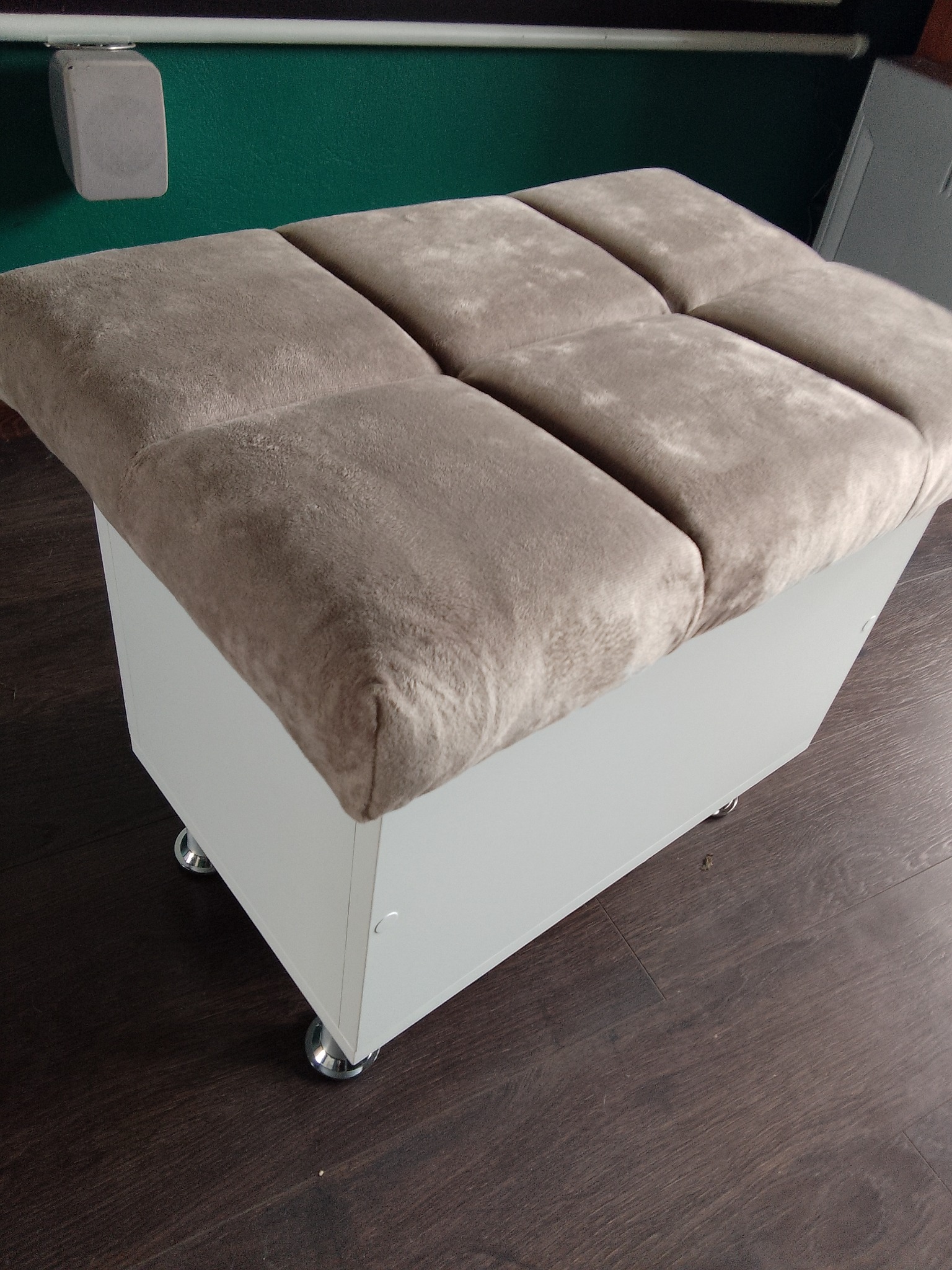 Pufa tapicerowana z beżowym siedziskiem podzielonym na sześć kwadratów, osadzona na białej skrzyni z kółkami, na tle ciemnej podłogi i zielonej ściany.