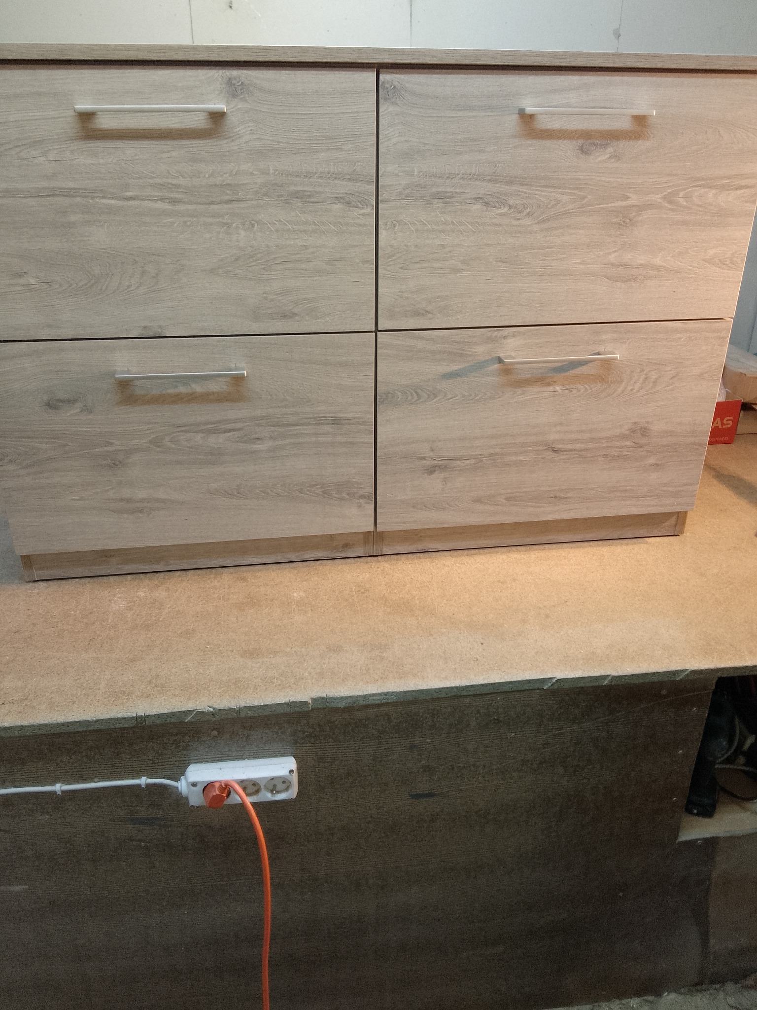 Cztery szuflady z jasnego drewna z poziomymi, srebrnymi uchwytami, umieszczone na blacie roboczym. Widoczny fragment przedłużacza i ciemne tło pod blatem.