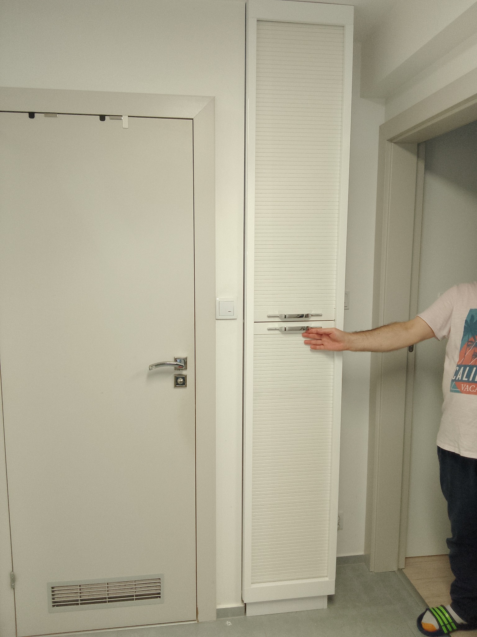 Biała szafa wnękowa z żaluzjowymi drzwiami, otwierana przez osobę. Widoczna obok biała ściana z włącznikiem i białe drzwi. Minimalistyczny design.