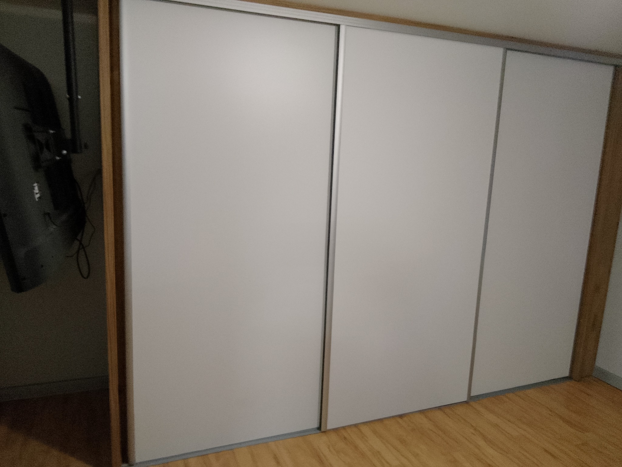 Biała szafa wnękowa z przesuwnymi drzwiami i drewnianą podłogą. Widoczny fragment telewizora wewnątrz szafy. Minimalistyczny design.