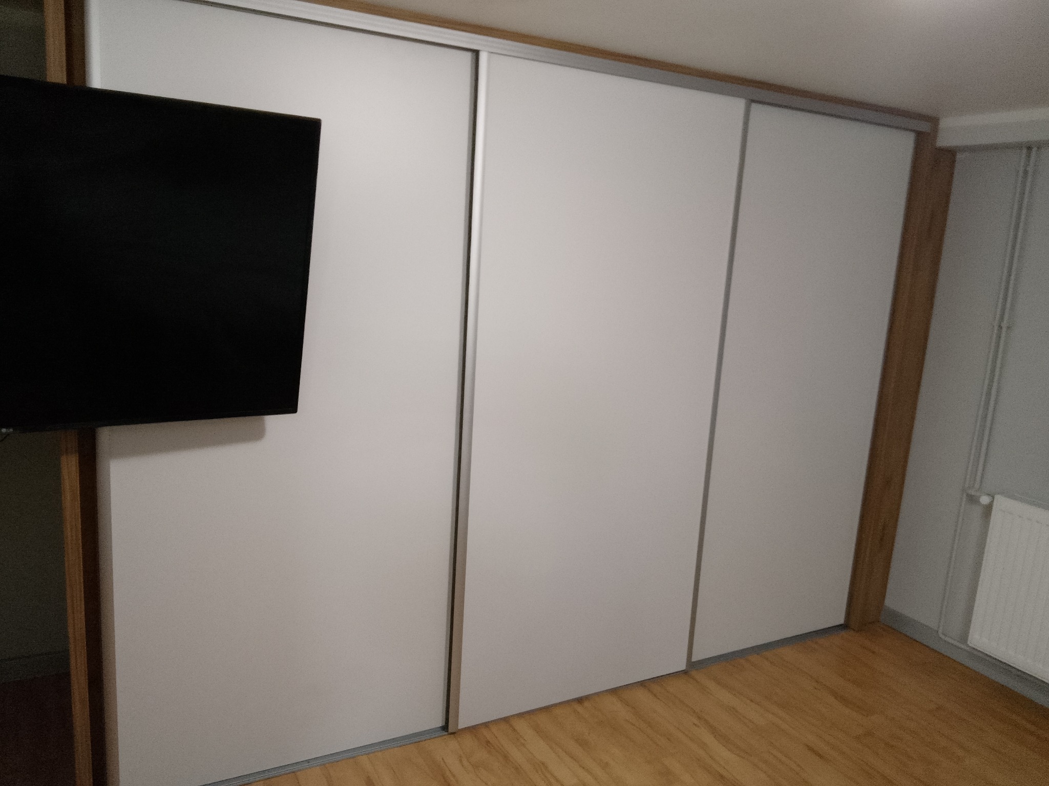 Biała szafa wnękowa z przesuwnymi drzwiami i telewizorem po lewej stronie. Widoczna drewniana podłoga i fragment grzejnika. Minimalistyczny design.