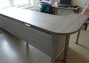 Szary, nowoczesny narożny stół biurowy z blatem imitującym drewno, na metalowych nogach. Widok z boku, z oknem i regałem w tle. Minimalistyczny design.