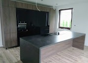 Nowoczesna kuchnia z wyspą: szare fronty szafek, czarny panel z piekarnikiem i mikrofalówką, minimalistyczny design, surowy stan wykończenia.