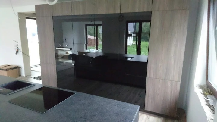 Nowoczesna kuchnia z wyspą i zabudową w odcieniach szarości i drewna. Widoczne okno i elementy wykończeniowe. Minimalistyczny design z czarnymi akcentami.