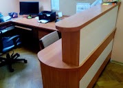 Recepcja z blatem w odcieniach drewna i białymi panelami, widoczna z boku, z biurkiem i sprzętem biurowym w tle. Ujęcie z niskiej perspektywy.