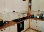 Jasna kuchnia w kształcie litery L z drewnianym blatem i czarną płytą grzewczą. Na półkach widoczne książki i akcesoria kuchenne. Ujęcie z bliska, naturalne światło.