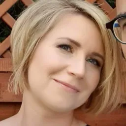 Portret uśmiechniętej blondynki z krótkimi włosami, na tle drewnianej konstrukcji i fragmentu osoby w okularach. Subtelny makijaż.