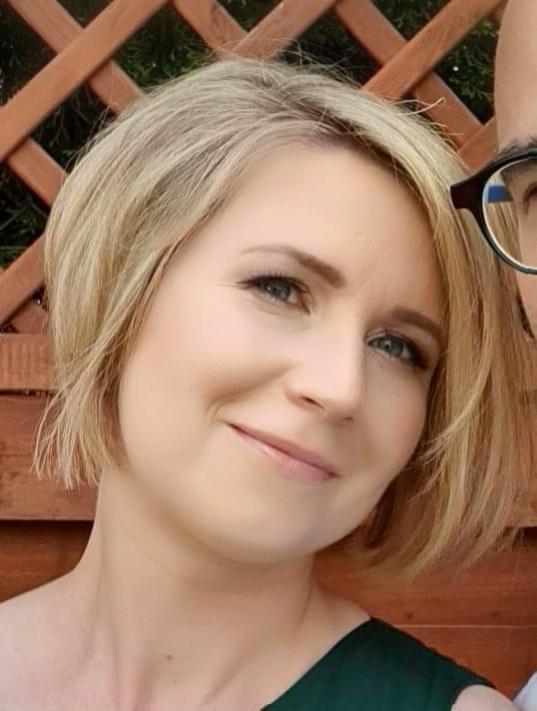 Portret uśmiechniętej kobiety z krótkimi blond włosami, wykonany w Piasecznie, z fragmentem okularów i drewnianej konstrukcji w tle.