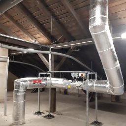 KAMENERGIA - Instalacja kanałów wentylacyjnych z izolacją termiczną na poddaszu, widoczne elementy mocujące i skrzynka sterownicza, oświetlenie punktowe, drewniana konstrukcja dachu.
