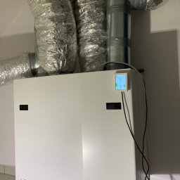KAMENERGIA - Biała centrala wentylacyjna z rekuperacją, widoczne aluminiowe rury wentylacyjne, zamontowany panel sterowania z kolorowym wyświetlaczem, szara podłoga z płytek.