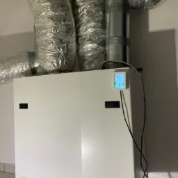 Biała centrala wentylacyjna z rekuperacją, widoczne aluminiowe rury wentylacyjne, zamontowany panel sterowania z kolorowym wyświetlaczem, szara podłoga z płytek.