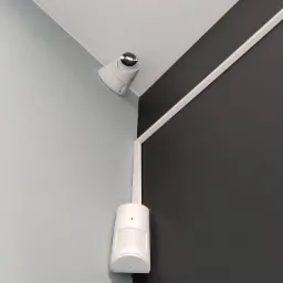 Kamera monitoringu kopułkowa i czujnik ruchu zamontowane w rogu pomieszczenia, ściany pomalowane na szaro i grafitowo, listwa przypodłogowa.