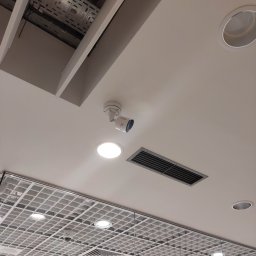 KAMENERGIA - Widok sufitu podwieszanego z kratownicą, oświetleniem LED, okrągłymi lampami halogenowymi, kratką wentylacyjną i kamerą przemysłową Hikvision.