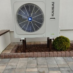 KAMENERGIA - Zewnętrzna jednostka pompy ciepła Kaisai na metalowych wspornikach, umieszczona na kostce brukowej obok rabaty z tujami, widok z przodu.
