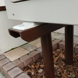 KAMENERGIA - Detale montażu zewnętrznej jednostki urządzenia grzewczego na brązowym stelażu, widoczne elementy mocujące i podłoże wyłożone kostką oraz korą.