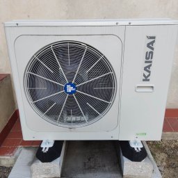 KAMENERGIA - Zewnętrzna jednostka klimatyzacji Kaisai zamontowana na betonowych blokach z czarnymi podkładkami antywibracyjnymi, widoczne logo 'We care about air' na osłonie wentylatora.