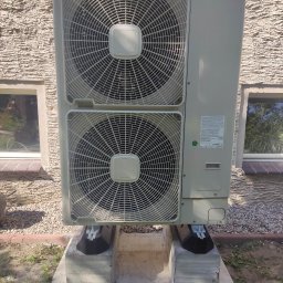 KAMENERGIA - Zewnętrzna jednostka klimatyzacyjna Hitachi zamontowana na betonowym fundamencie z amortyzatorami, na tle elewacji budynku z fakturą baranka i widocznymi oknami.