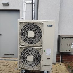 KAMENERGIA - Zewnętrzna jednostka dwuwentylatorowej pompy ciepła Hitachi, zamontowana na betonowych podstawach przy szarej ścianie budynku z widoczną instalacją rurową i skrzynką na listy.