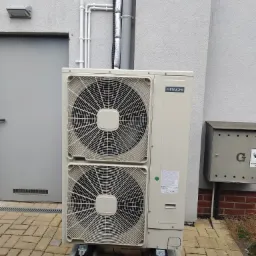 Zewnętrzna jednostka dwuwentylatorowej pompy ciepła Hitachi, zamontowana na betonowych podstawach przy szarej ścianie budynku z widoczną instalacją rurową i skrzynką na listy.