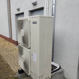 KAMENERGIA - Zewnętrzna jednostka klimatyzacyjna Hitachi, zamontowana na betonowej podstawie przy szarej ścianie budynku z widocznymi rurami instalacyjnymi i kostką brukową.