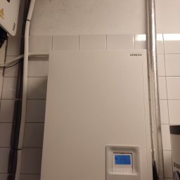 KAMENERGIA - Biała pompa ciepła Hitachi zamontowana na ścianie z białych płytek, z widocznymi rurami w izolacji i panelem sterowania z niebieskim wyświetlaczem.