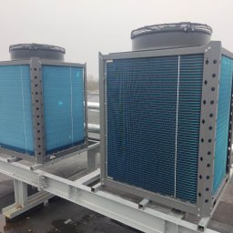 KAMENERGIA - Dwa agregaty chłodnicze z wentylatorami na dachu budynku, zamontowane na stalowej konstrukcji wsporczej. Widoczny również klimatyzator okienny i komin wentylacyjny.