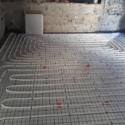 Rozłożone białe rury ogrzewania podłogowego na czarnej siatce z pomarańczowymi znacznikami, widoczne surowe ściany w tle.