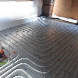 KAMENERGIA - Rozłożone rury ogrzewania podłogowego w surowym stanie, widoczne narzędzia i elementy montażowe, ściany w trakcie wykończenia.