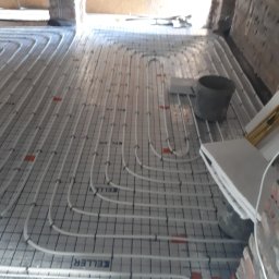 KAMENERGIA - Rozłożone rury ogrzewania podłogowego na folii z siatką, widoczny betonowy kubeł i fragmenty styropianu, mur w tle.