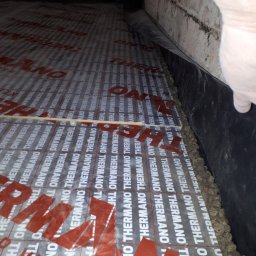 KAMENERGIA - Montaż izolacji podłogowej z folii z nadrukiem 'THERMANO' i warstwy keramzytu przy ścianie z cegły, widoczne rękawice robocze.