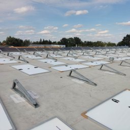 KAMENERGIA - Montaż paneli fotowoltaicznych na płaskim dachu budynku, widoczne metalowe stelaże i częściowo ułożone panele z przewodami, w tle drzewa i błękitne niebo z chmurami.