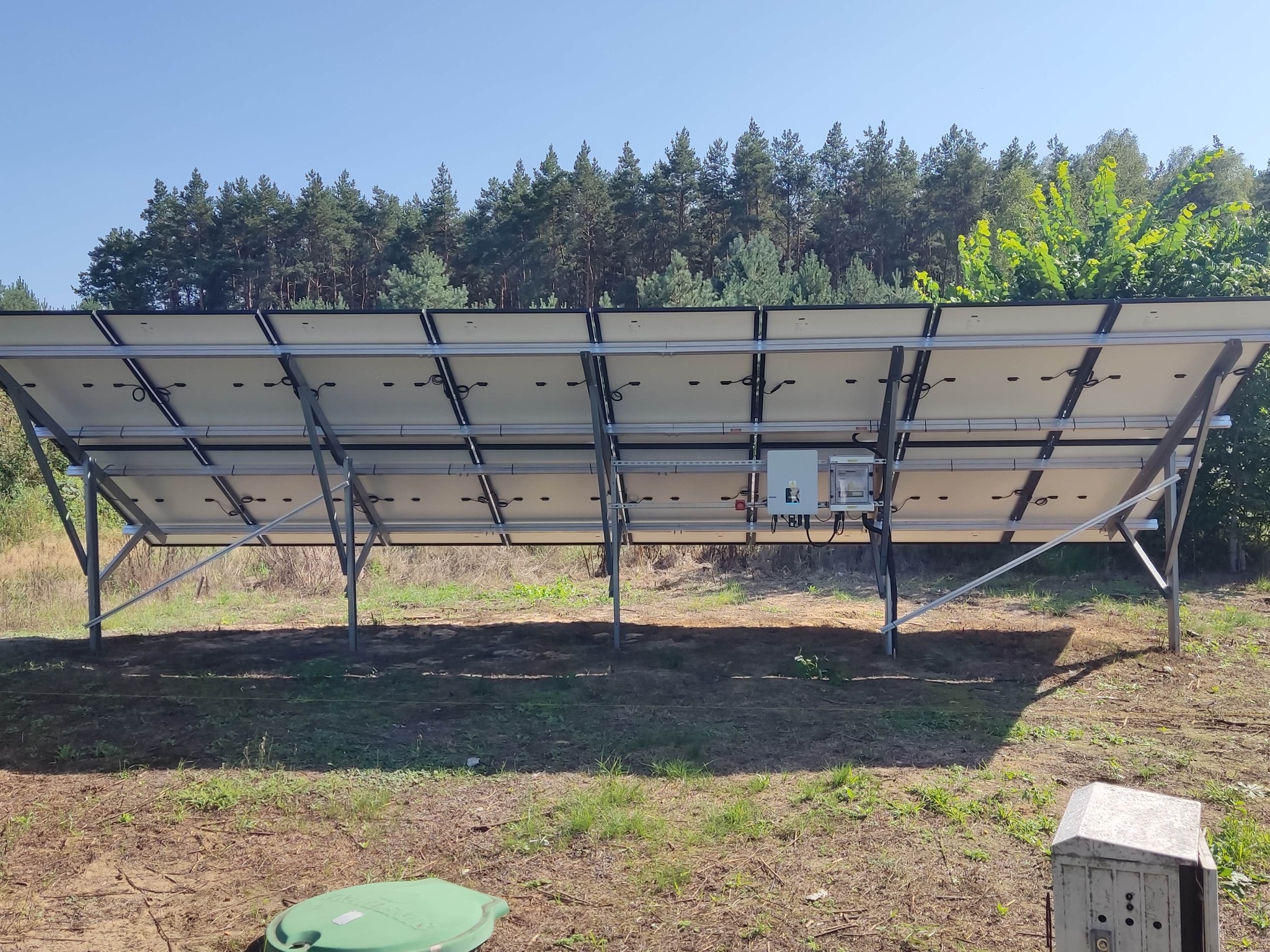 Widok od spodu na instalację paneli słonecznych we Wrocławiu, z widocznymi metalowymi wspornikami i okablowaniem, na tle zielonego lasu i błękitnego nieba.