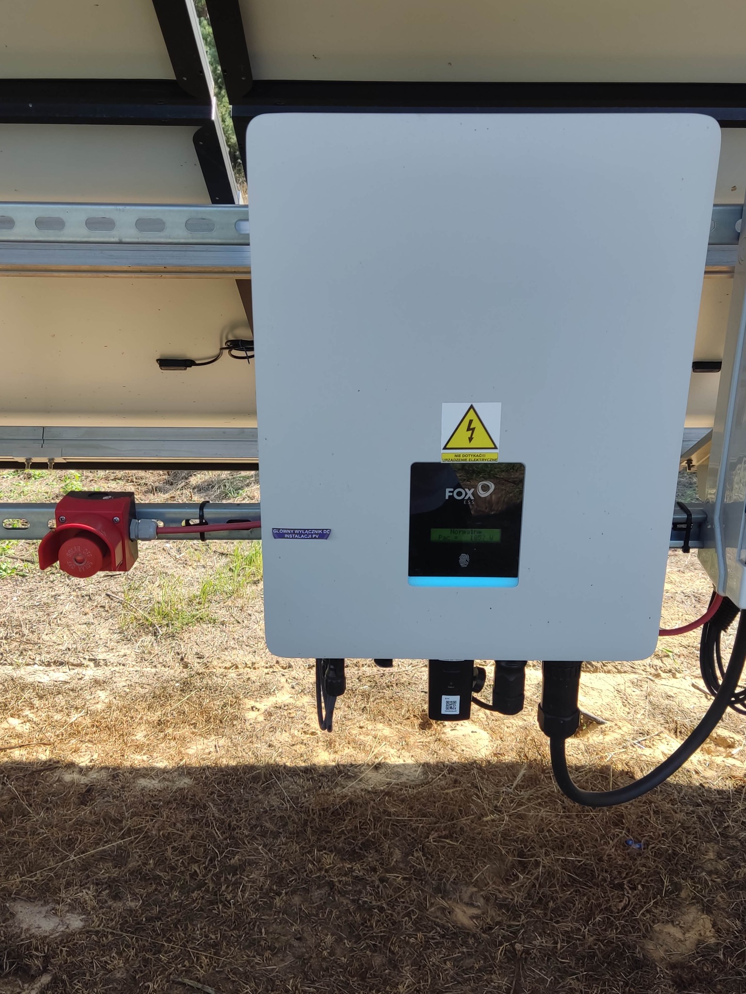 Szary inwerter fotowoltaiczny marki FOX ESS z widocznym wyświetlaczem i ostrzeżeniem o wysokim napięciu, zamontowany na konstrukcji z paneli słonecznych, z czerwonym wyłącznikiem bezpieczeństwa po...