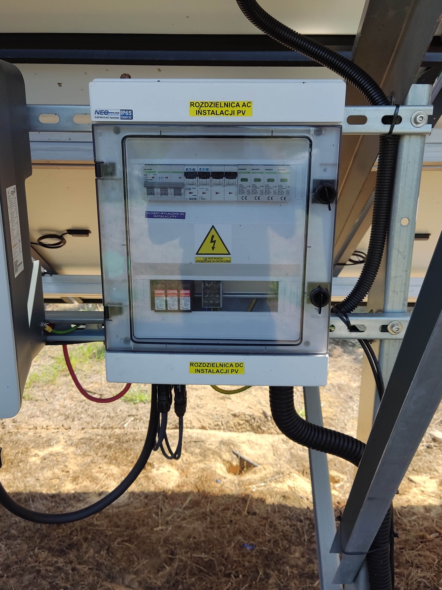 Rozdzielnica AC/DC instalacji fotowoltaicznej zamontowana na konstrukcji wsporczej, widoczne zabezpieczenia, wyłączniki, przewody oraz ostrzeżenie o napięciu.