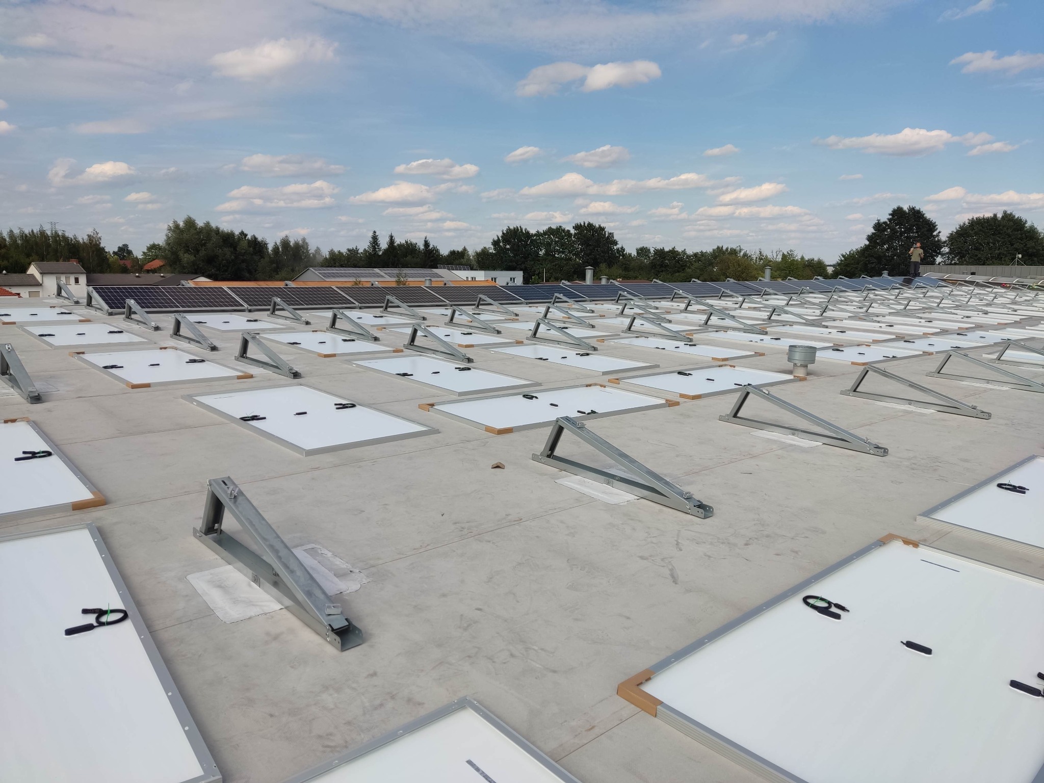 Montaż paneli fotowoltaicznych na płaskim dachu budynku, widoczne metalowe stelaże i częściowo ułożone panele z przewodami, w tle drzewa i błękitne niebo z chmurami.
