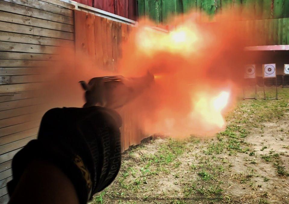 Dynamiczne ujęcie wystrzału z rewolweru z efektem pomarańczowego dymu na tle drewnianej ściany i tarcz strzeleckich.