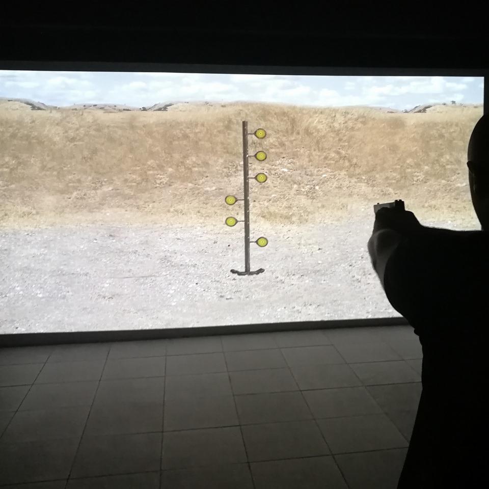Strzelanie z pistoletu do tarczy z żółtymi okręgami na tle wyświetlanego krajobrazu polnego, w pomieszczeniu ze szarą podłogą.