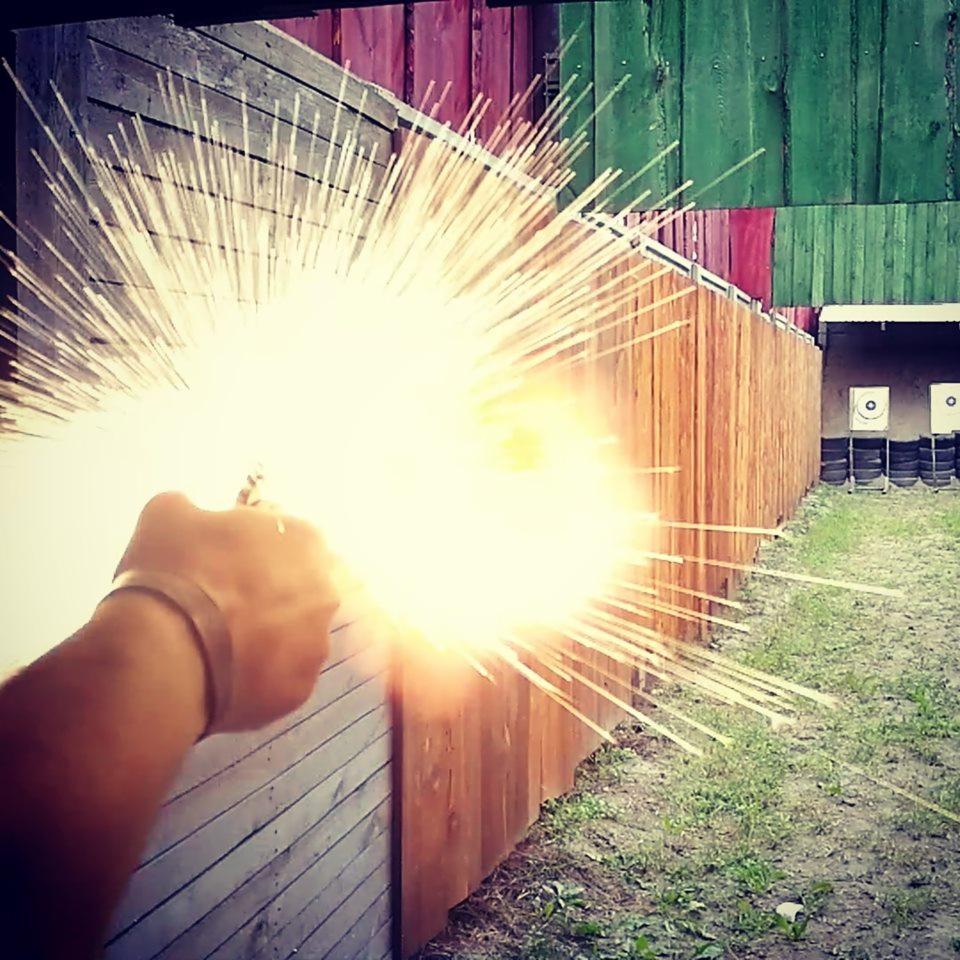 Moment wystrzału z pistoletu na strzelnicy, widoczna ręka, błysk i tarcze w tle.