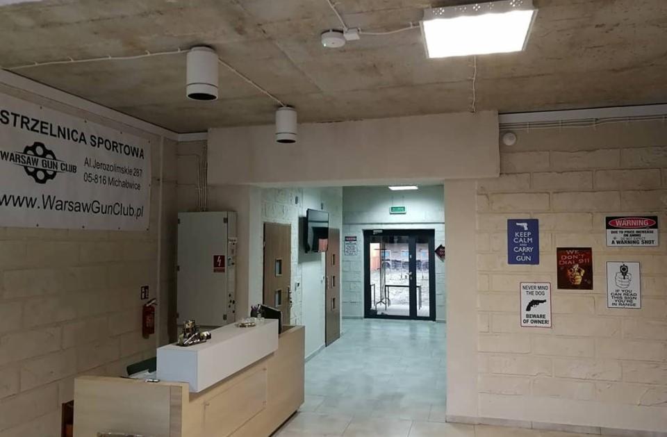 Wnętrze strzelnicy sportowej z recepcją, widocznymi plakatami z motywami broni i zasad bezpieczeństwa oraz logo Warsaw Gun Club