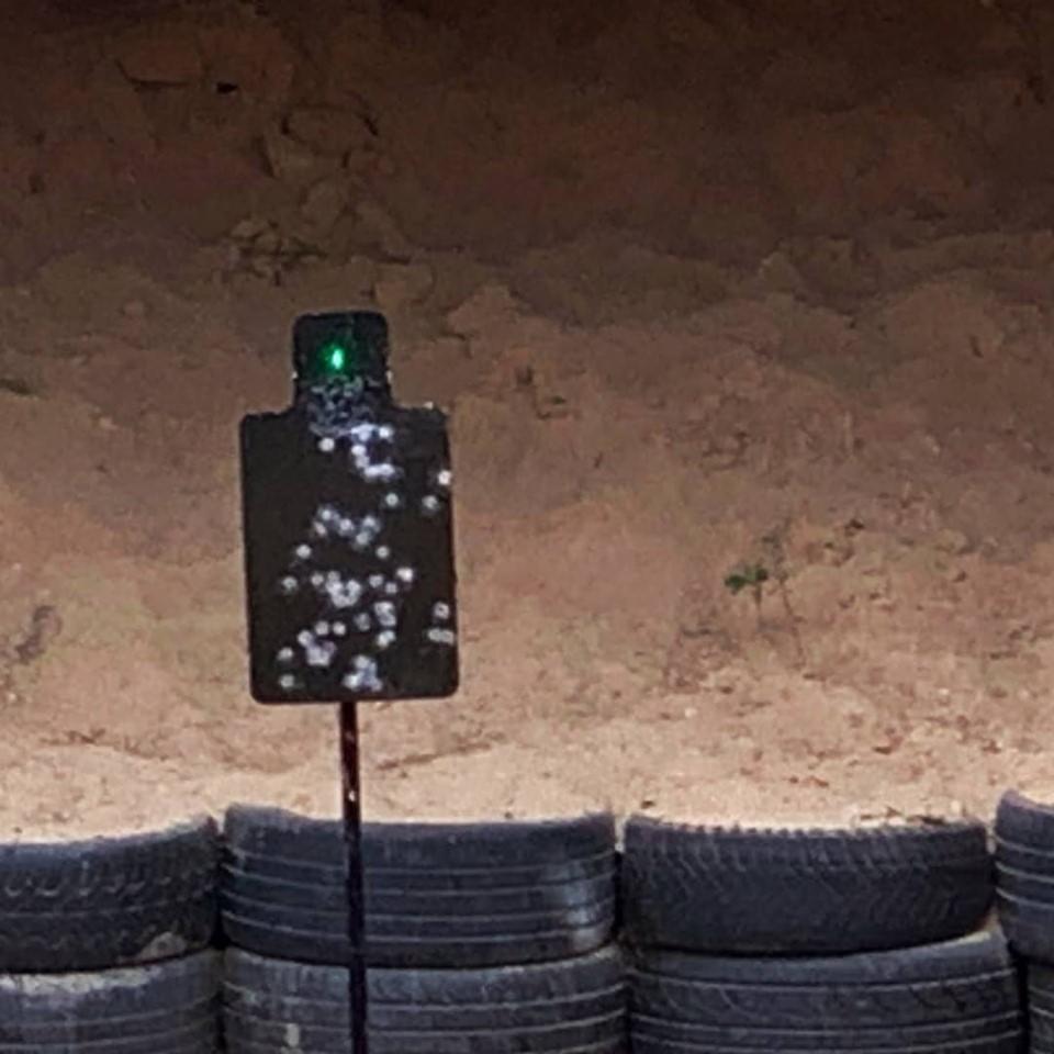 Tarcza strzelecka z licznymi śladami po kulach i zielonym laserowym wskaźnikiem, ustawiona na tle ziemnego wału i zabezpieczenia z opon.